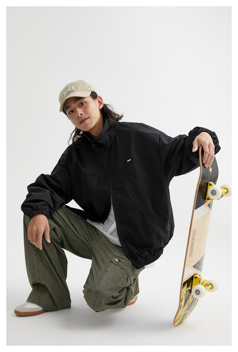 Model crouching in a black zip-front jacket with elastic cuffs and hem, beside a skateboard.