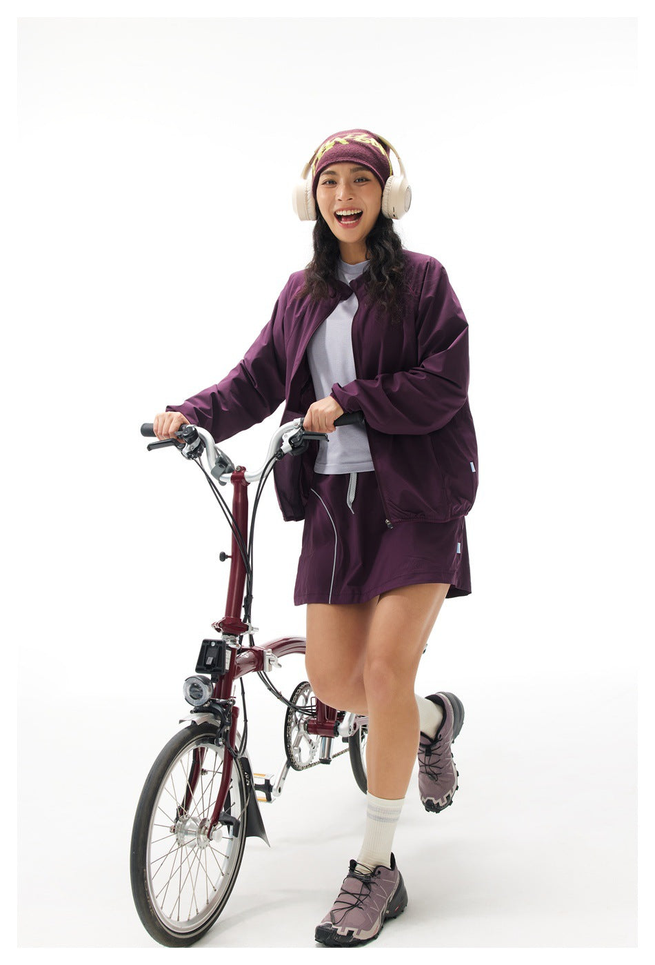 Model wearing a plum windbreaker and matching shorts while holding a bicycle; yellow graphic on her headband.