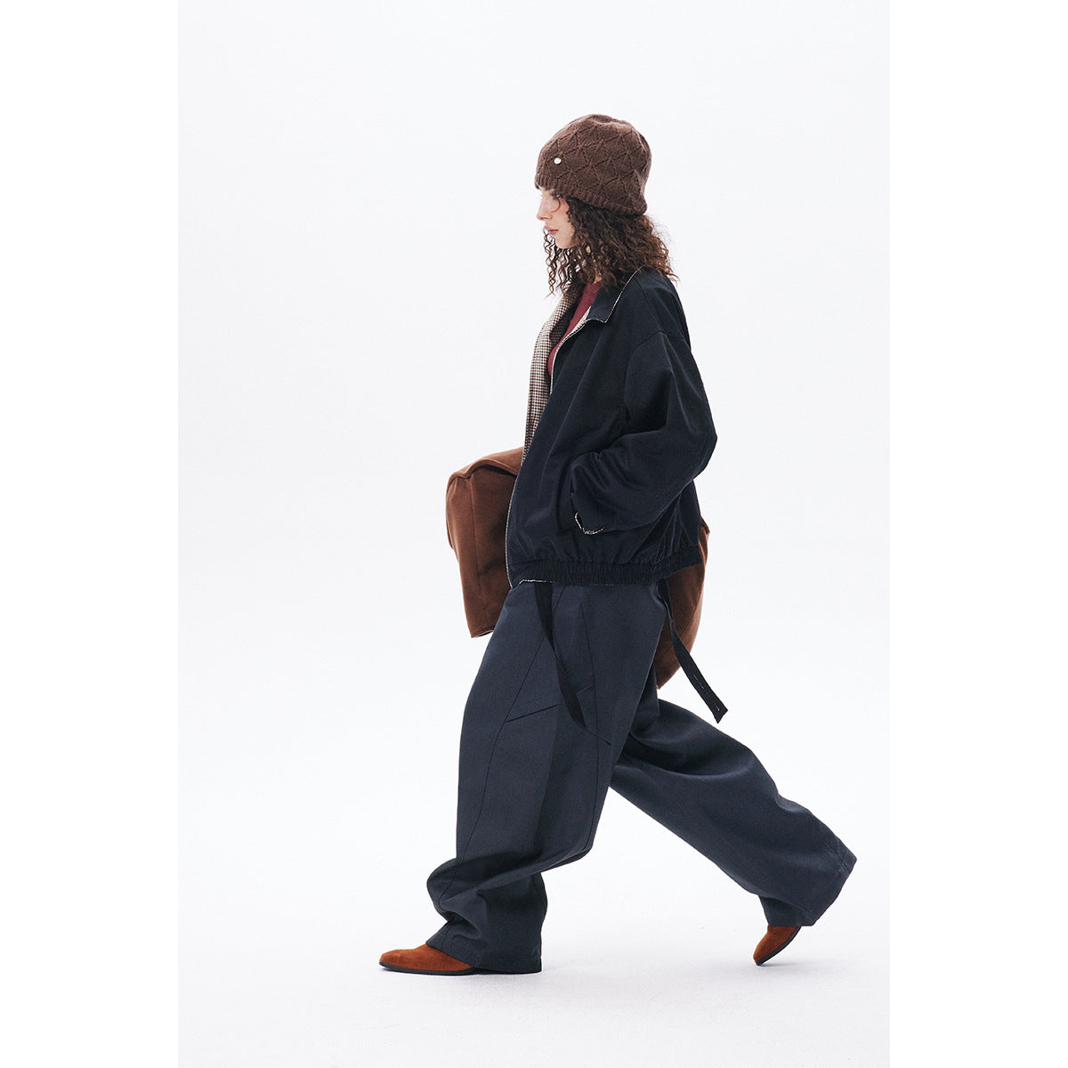 Side view of a model wearing a dark navy jacket and loose trousers, with a brown knit beanie and rust-colored bag.