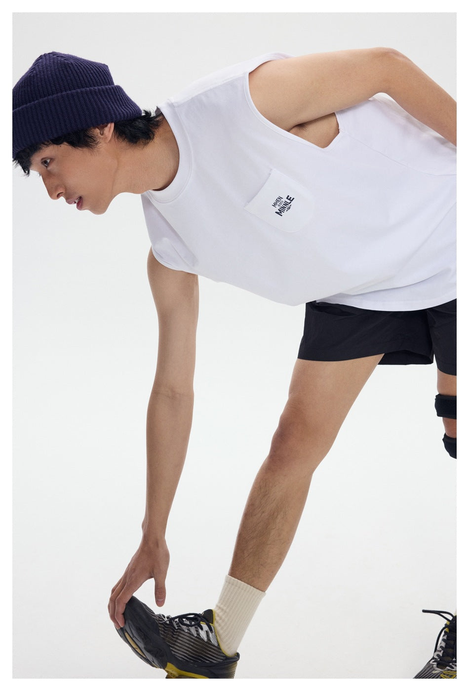 Model in a white sleeveless shirt with a small printed chest pocket, black shorts, and a navy beanie.