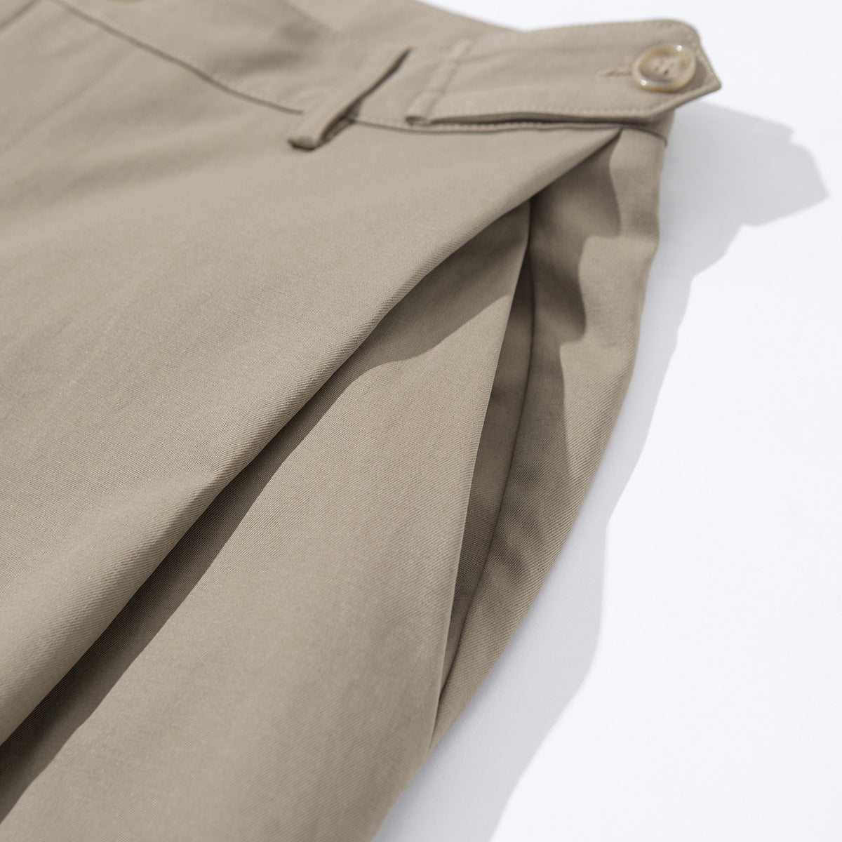 Close-up of taupe trousers showing a buttoned waist tab and folded fabric.