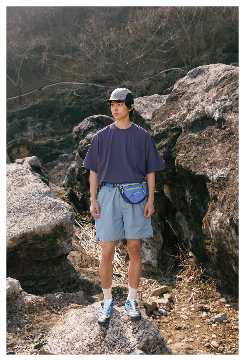 Full-length model in a purple pocket T-shirt and light blue shorts with a waist bag outdoors.