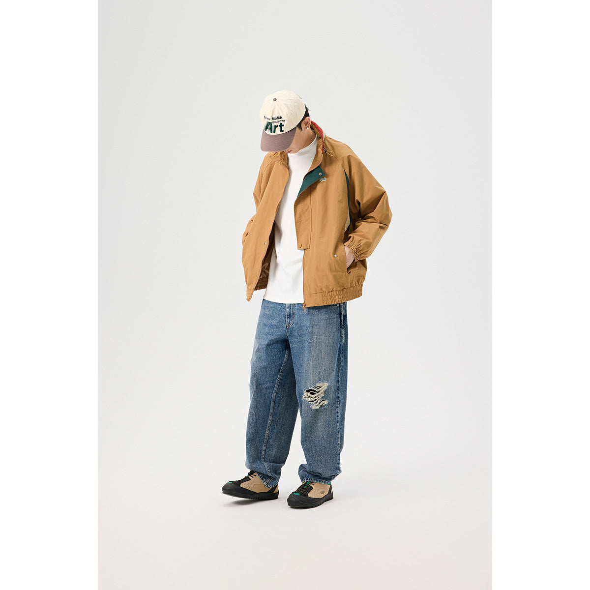 Model wearing a tan hooded zip jacket with dark teal accents, white shirt, blue distressed jeans and a cap.