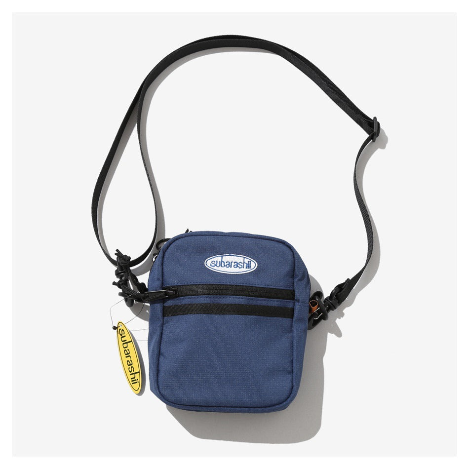 Blue crossbody bag with two black front zipper strips, oval logo patch and black shoulder strap
