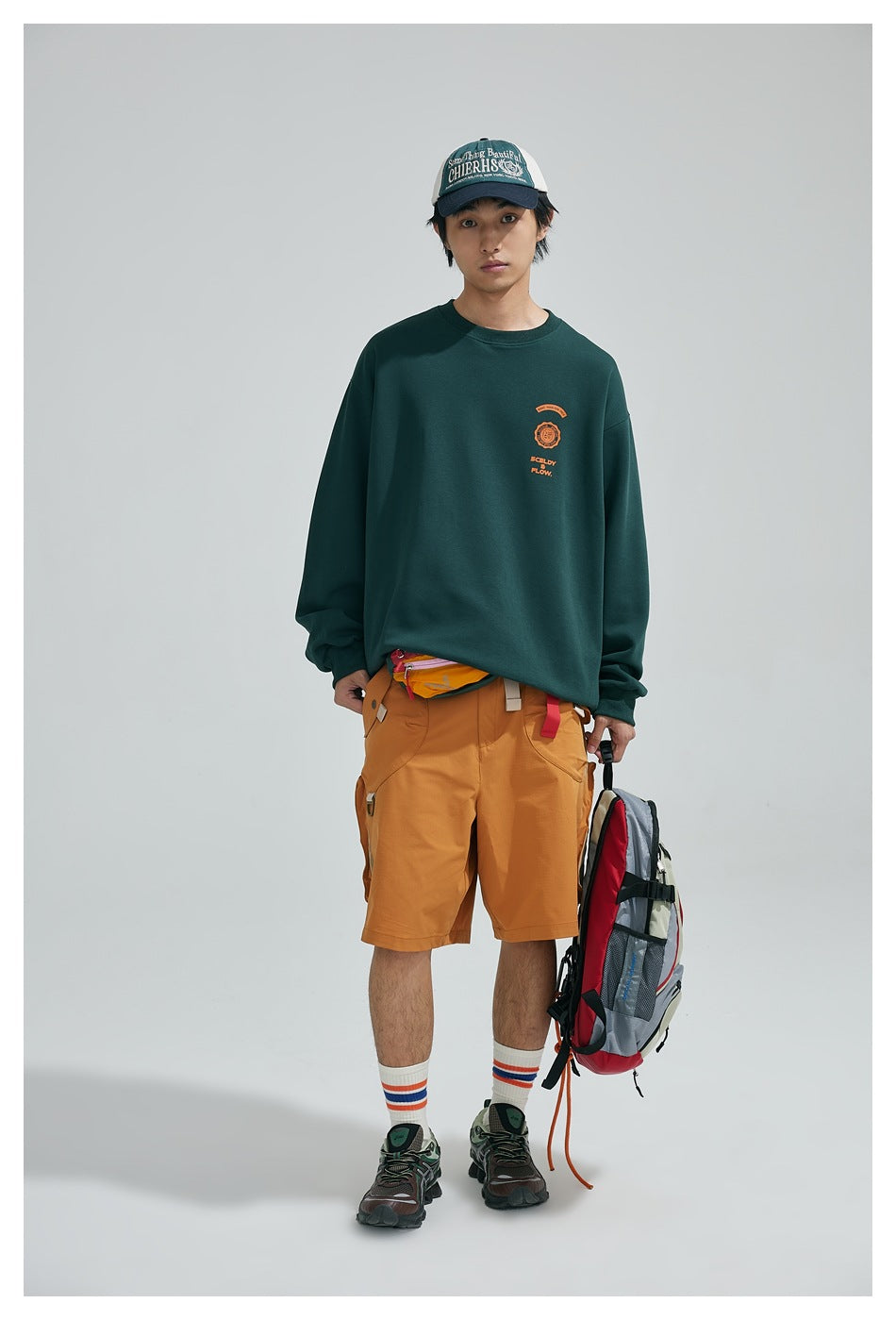 Model wearing a dark green crewneck sweatshirt with a small orange chest graphic, mustard cargo shorts, cap and backpack.