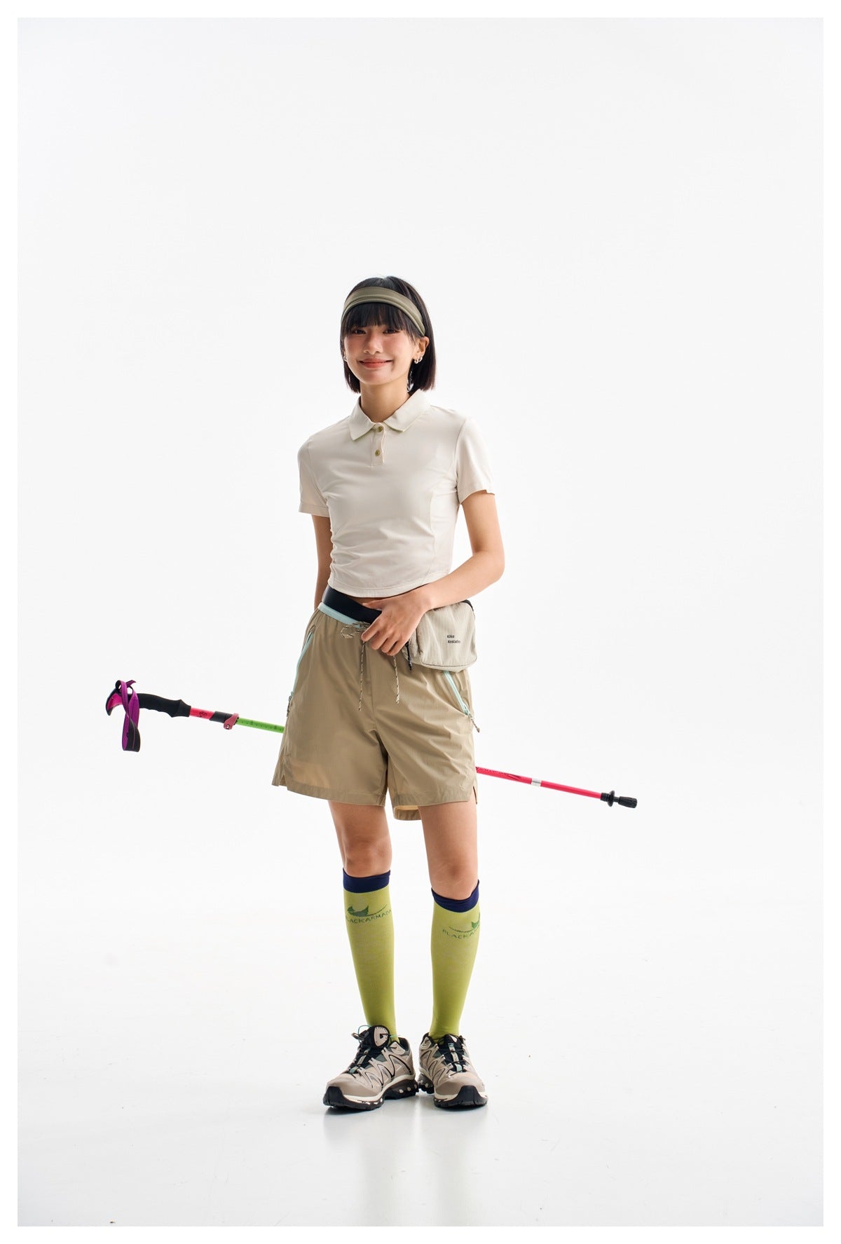 Model wearing a white cropped polo and khaki shorts, holding trekking poles against a white studio background.