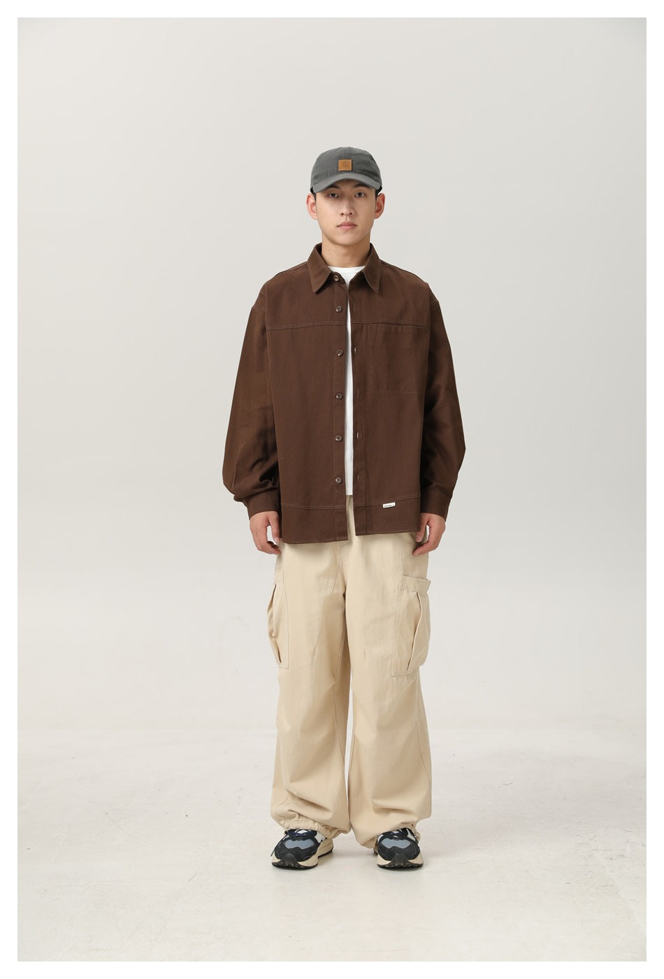 Man wearing a brown button-up shirt open over a white tee with beige cargo pants and a gray cap, front view