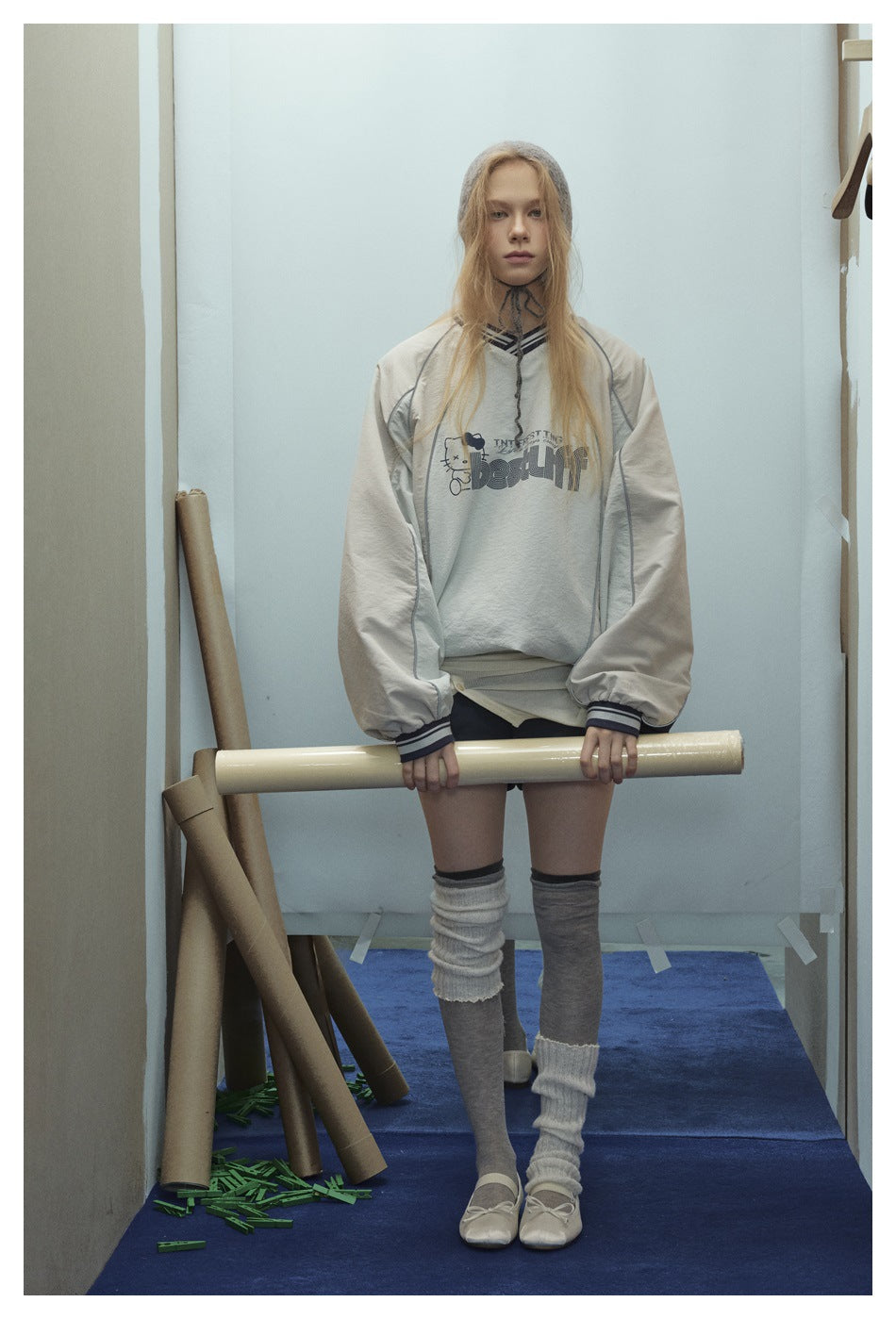 Model wearing an oversized light gray V-neck pullover with striped trim and chest graphic, holding a cardboard tube