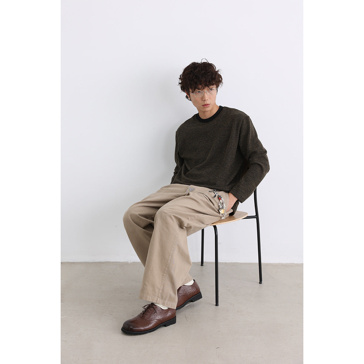 Seated model wearing a dark brown and black textured crewneck sweater with loose beige trousers.