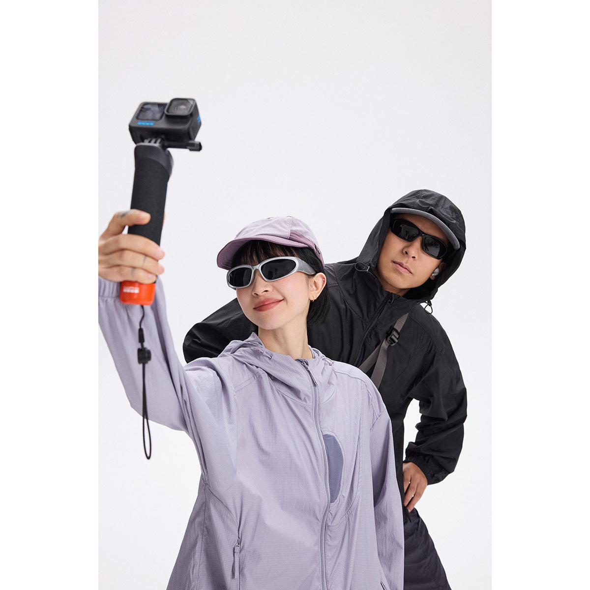 Two models pose in lavender and black hooded windbreakers; the woman holds an action camera on a grip.