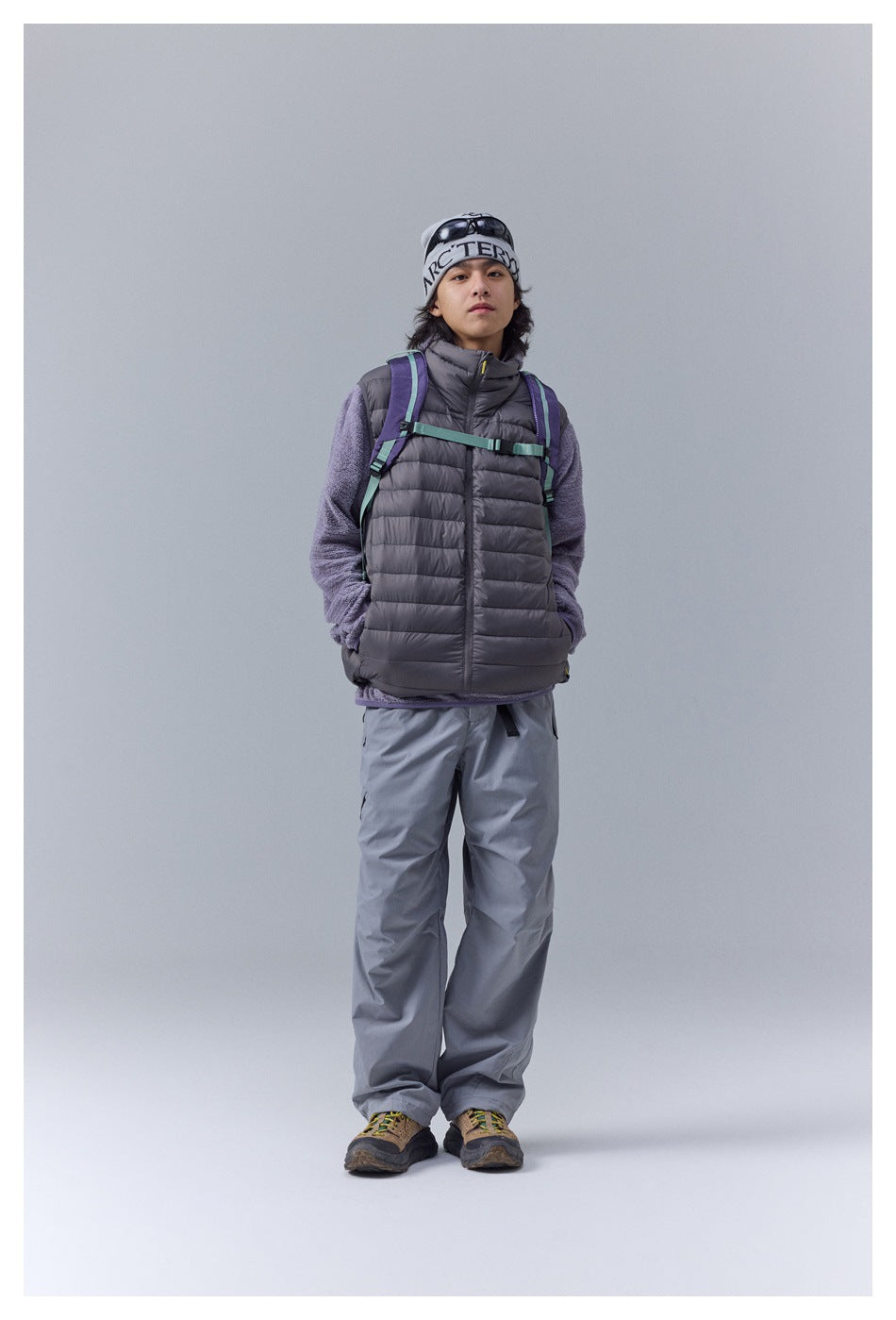 Model wearing a charcoal quilted puffer vest over a lavender fleece, with gray trousers and a backpack.