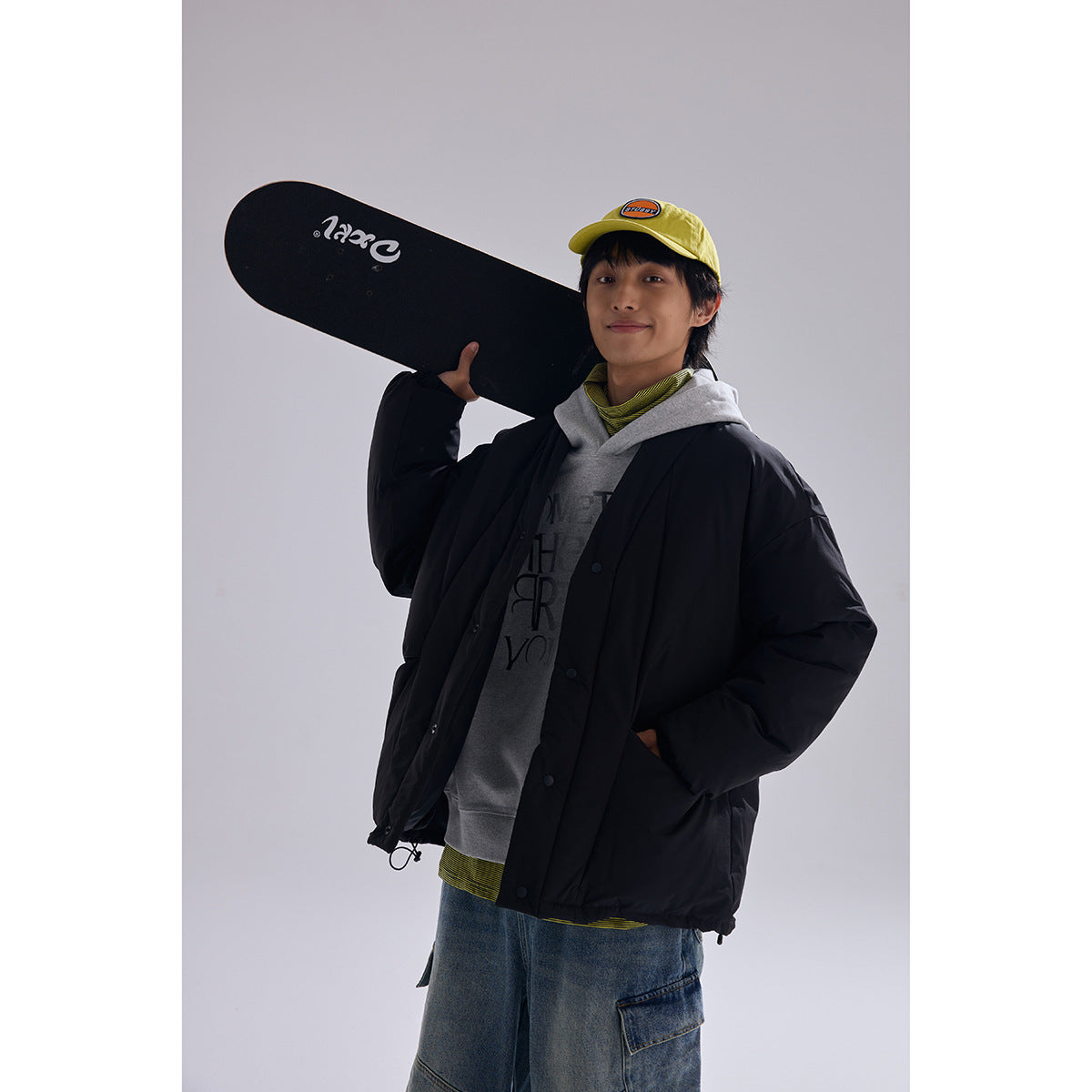 Model wearing a black oversized quilted jacket over a gray hoodie, holding a skateboard on one shoulder.