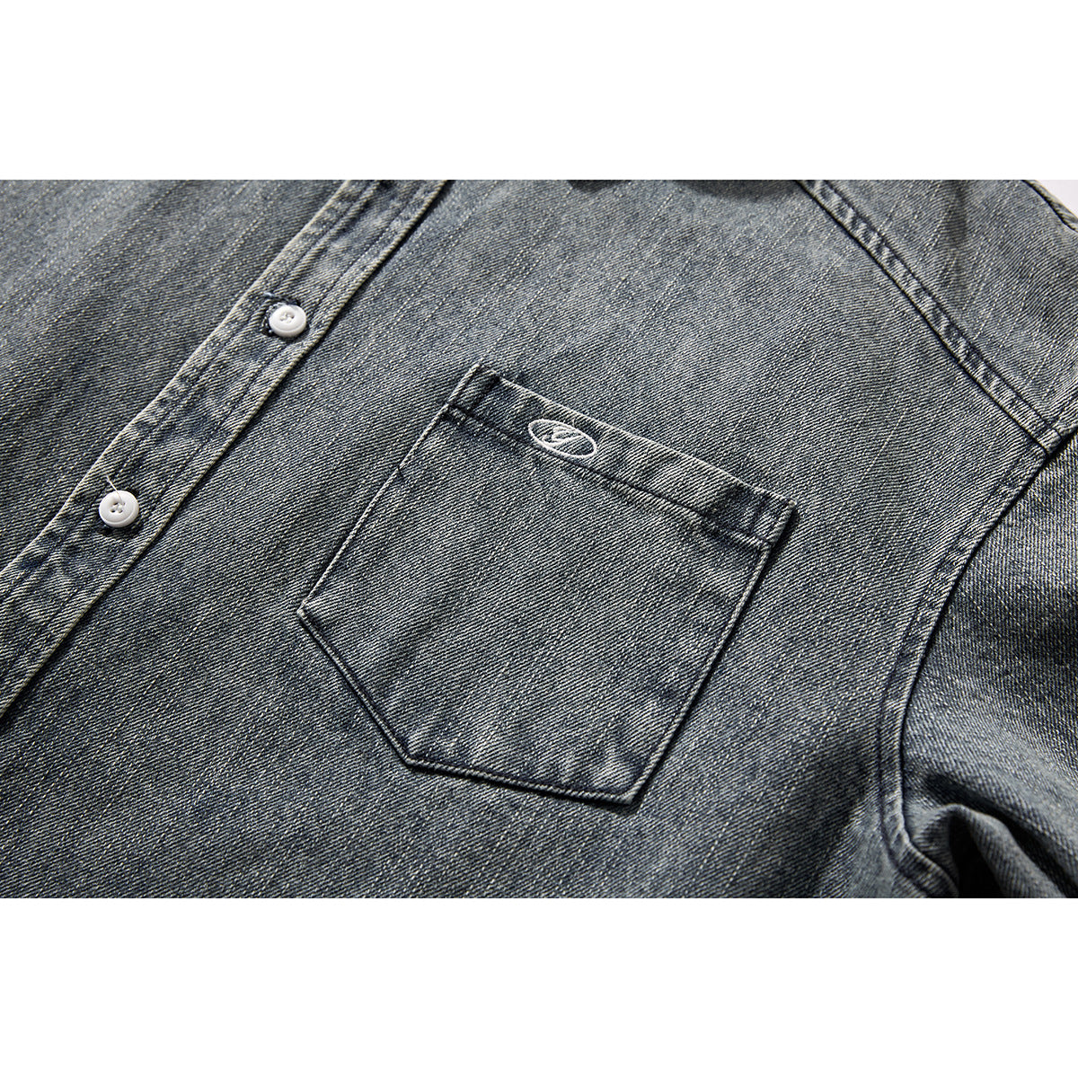 Close-up of a dark gray washed denim shirt chest pocket with a small embroidered oval and white buttons.