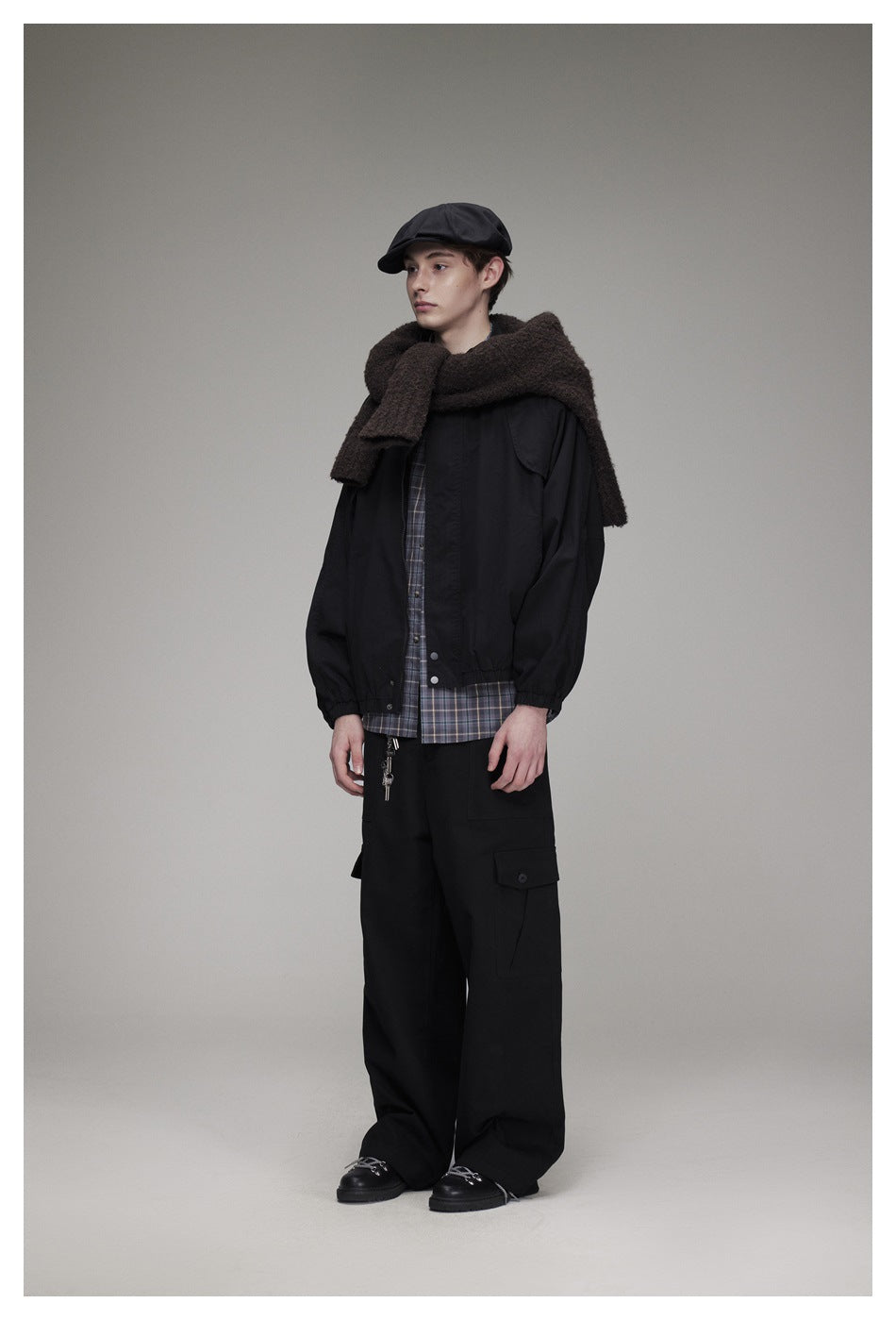 Model wearing a black jacket over a plaid shirt, wide black cargo trousers, a brown scarf and black cap.
