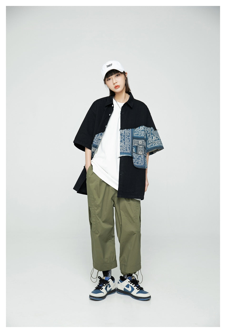 Model wearing black short-sleeve patchwork bandana shirt open over a white tee with olive pants and a white cap