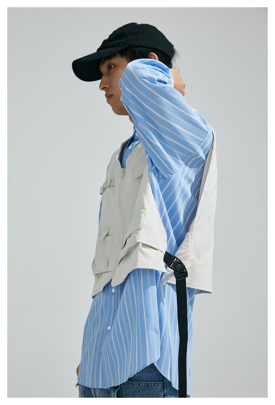 Person wearing an off-white utility vest over a blue striped shirt, shown from the side with a black cap.