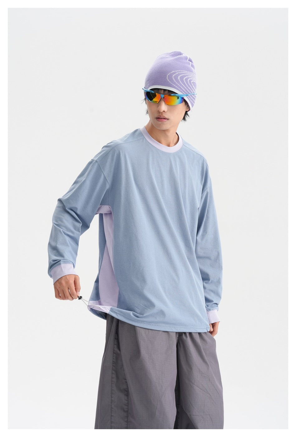Model wearing an oversized light blue long-sleeve shirt with lavender trim and side panels, pulling the hem outward.