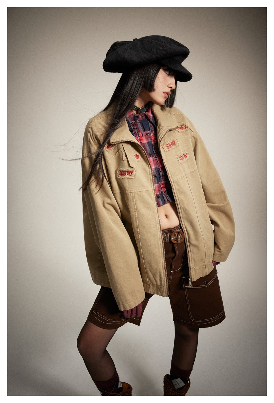 Model wearing a tan patch-decorated zip jacket over a red plaid shirt, dark brown shorts, and a black cap.