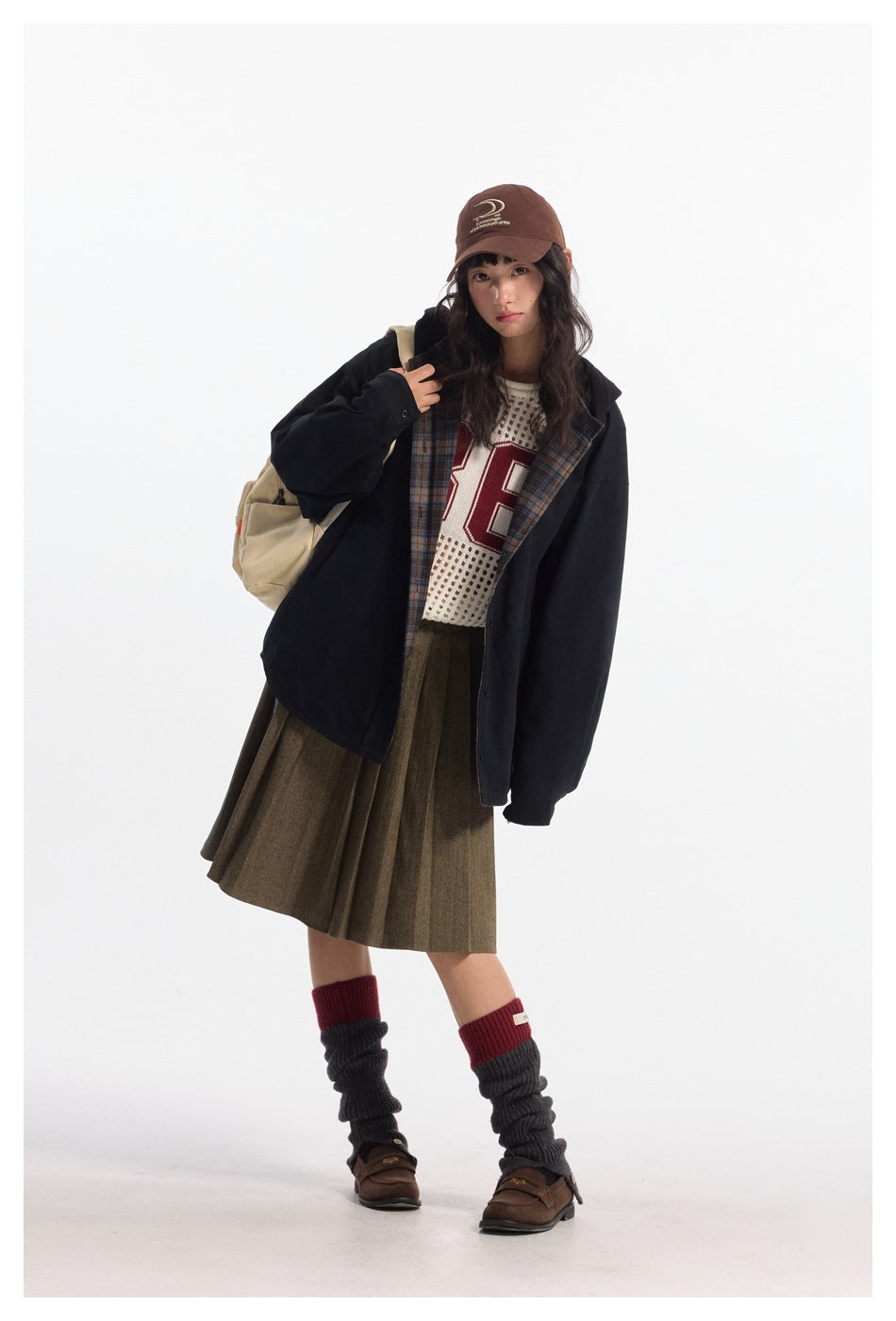 Model wearing an open black jacket with plaid lining over a cream mesh top and olive pleated skirt.
