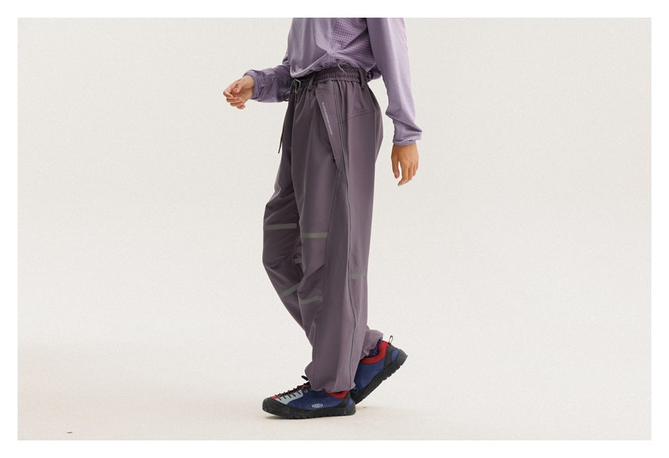 Cropped model wearing loose charcoal-purple pants with muted reflective bands, an elastic drawstring waist and gathered