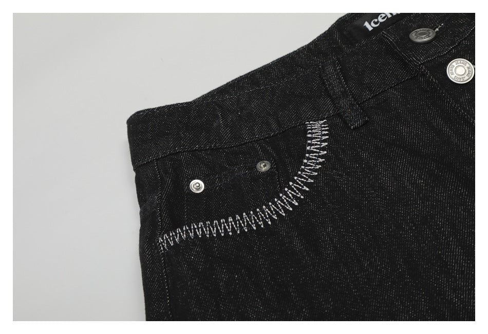 Close-up of black jeans with white zigzag stitching along a pocket edge, metal rivets, and waistband hardware.