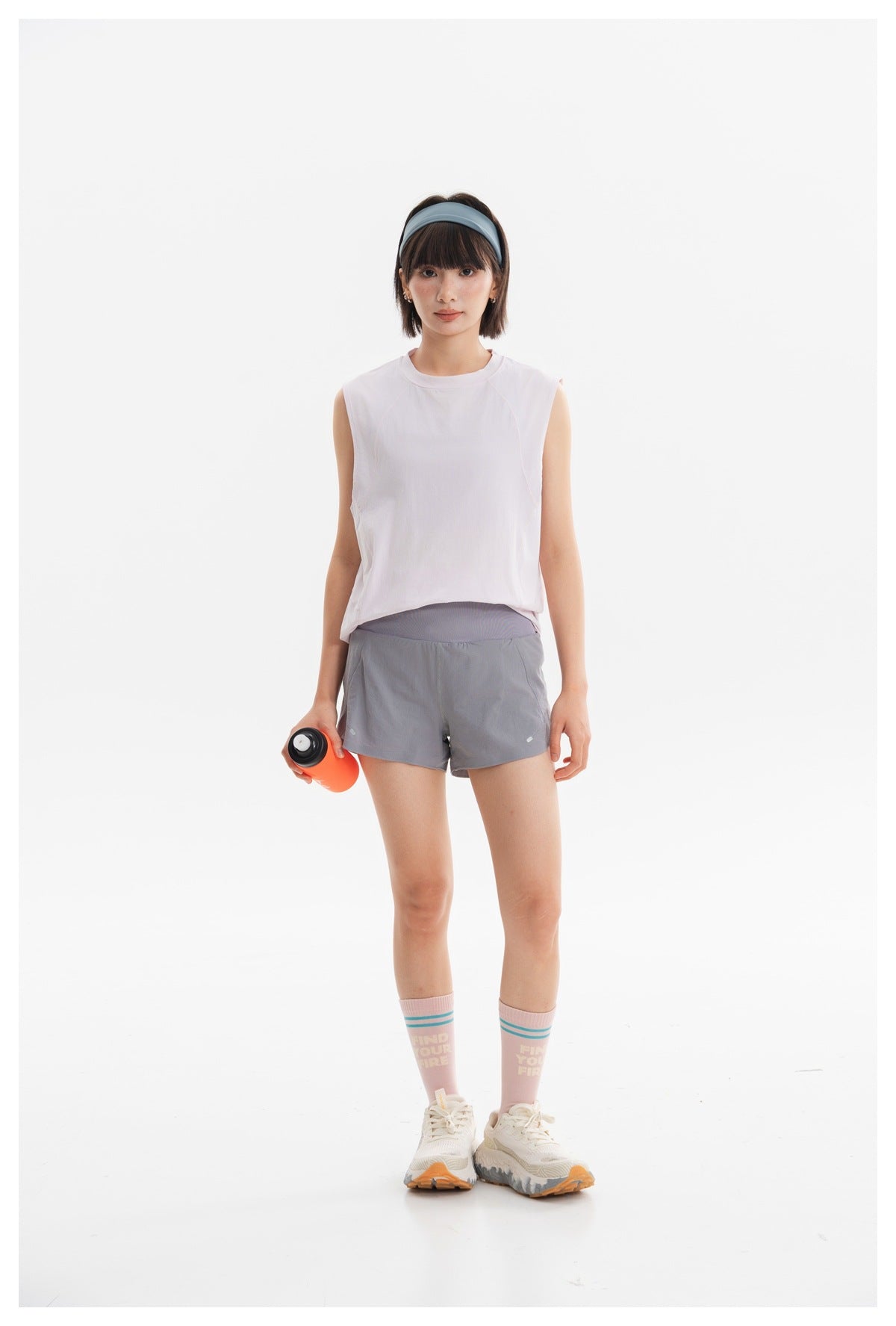 Woman wearing a white sleeveless top and gray athletic shorts, holding an orange bottle against a white background.