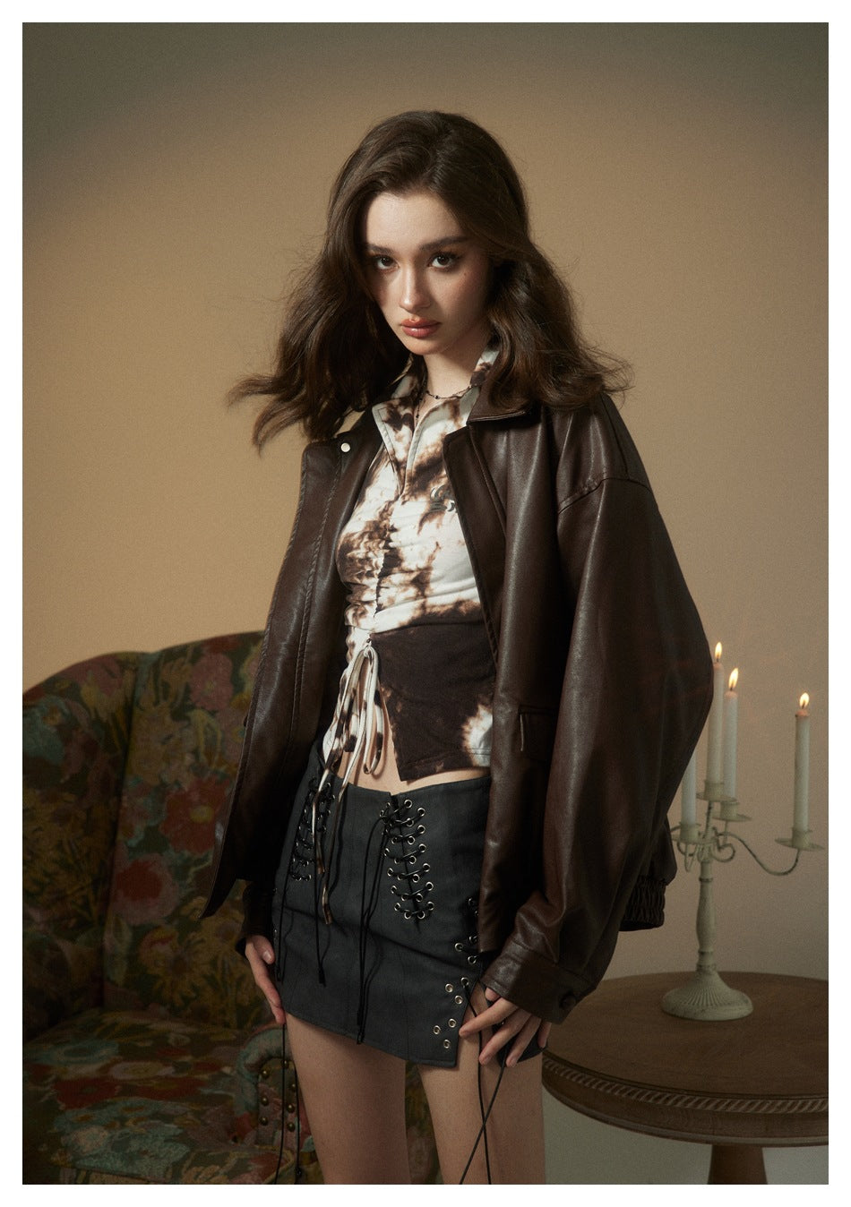 Model wearing an oversized dark brown leather-look jacket over a printed top and black lace-up mini skirt.