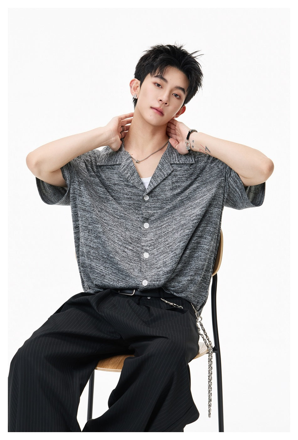 Seated model wearing a dark gray marled short-sleeve button-up shirt with a wide open collar and white buttons.