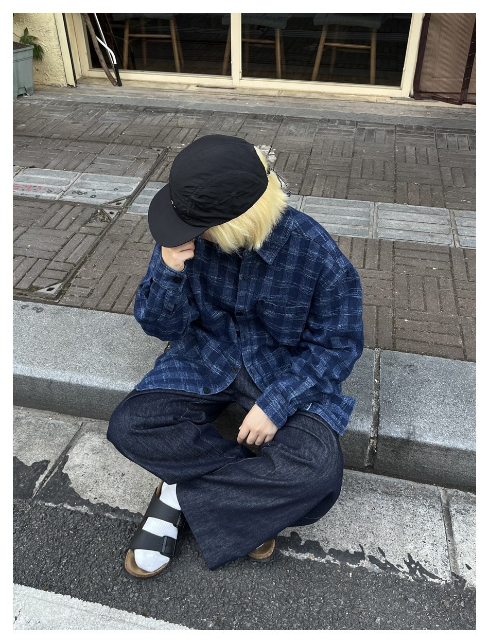 Person seated on a curb wearing the washed blue plaid shirt open over wide dark jeans, with a black cap