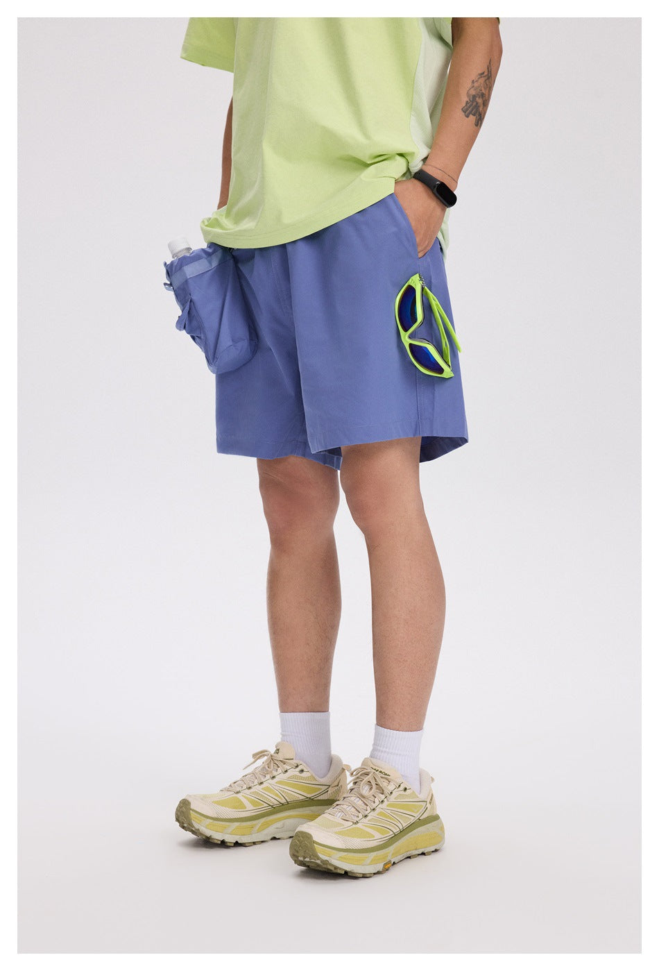 Angled view of a model in periwinkle-blue shorts with utility pockets and neon-green sunglasses clipped at the side.