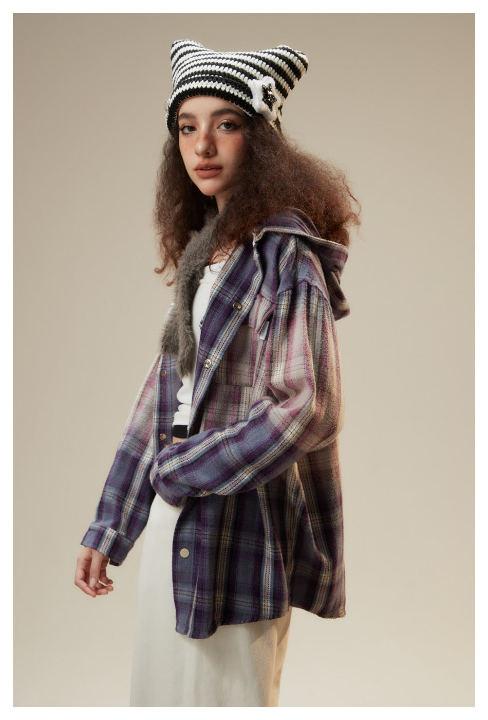 Three-quarter view of a model wearing a long purple plaid jacket with a hood and a black-and-white striped knit hat.