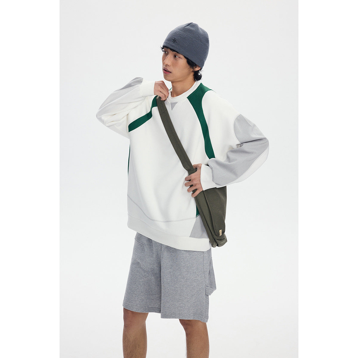 Model wearing a white sweatshirt with dark green and gray panels and gray shorts, carrying an olive crossbody bag.