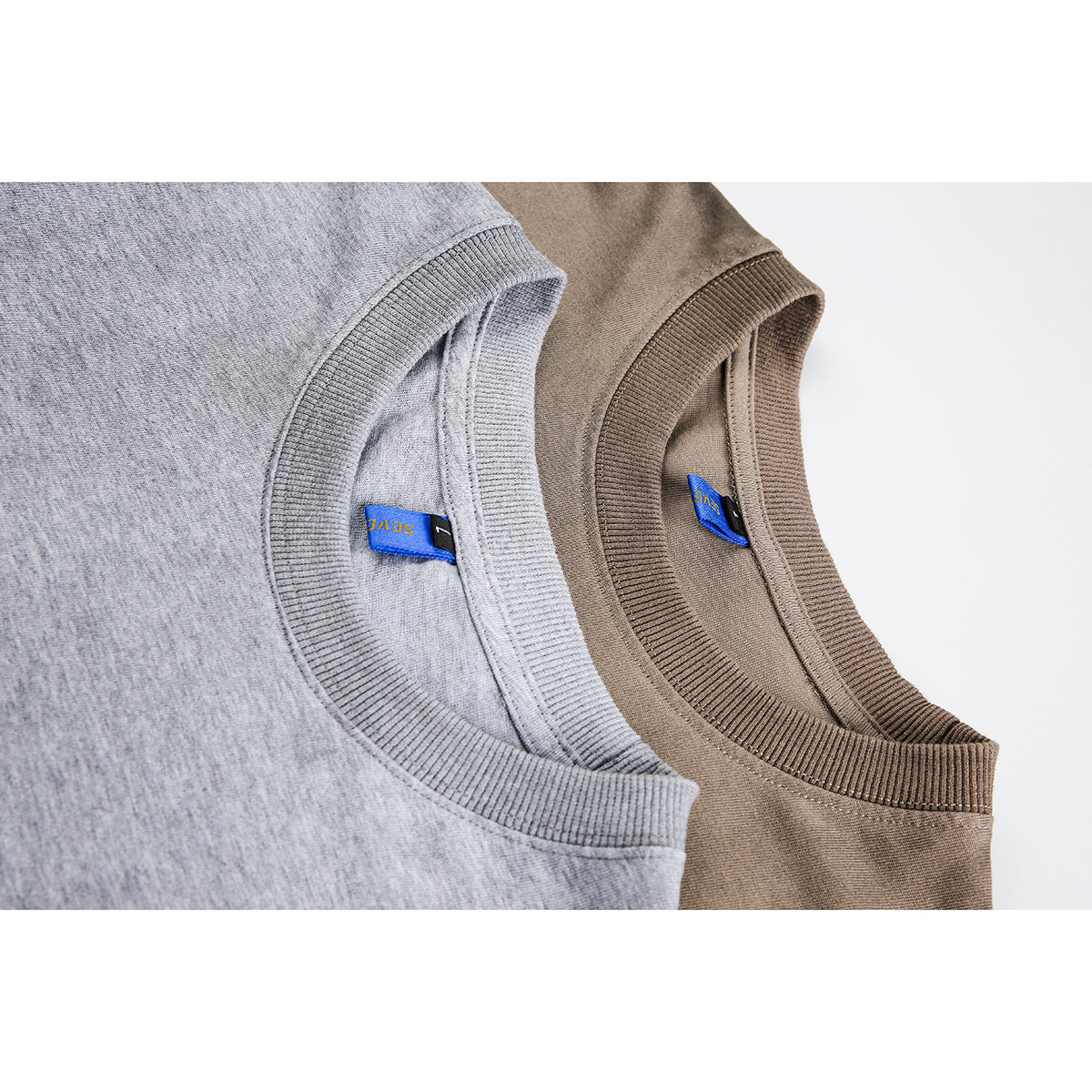 Close-up of the ribbed crew necklines and blue labels on light gray and taupe T-shirts.