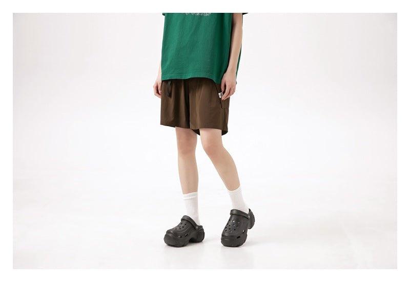 Cropped model wearing brown shorts with a green T-shirt, white socks, and black clogs; front three-quarter view.