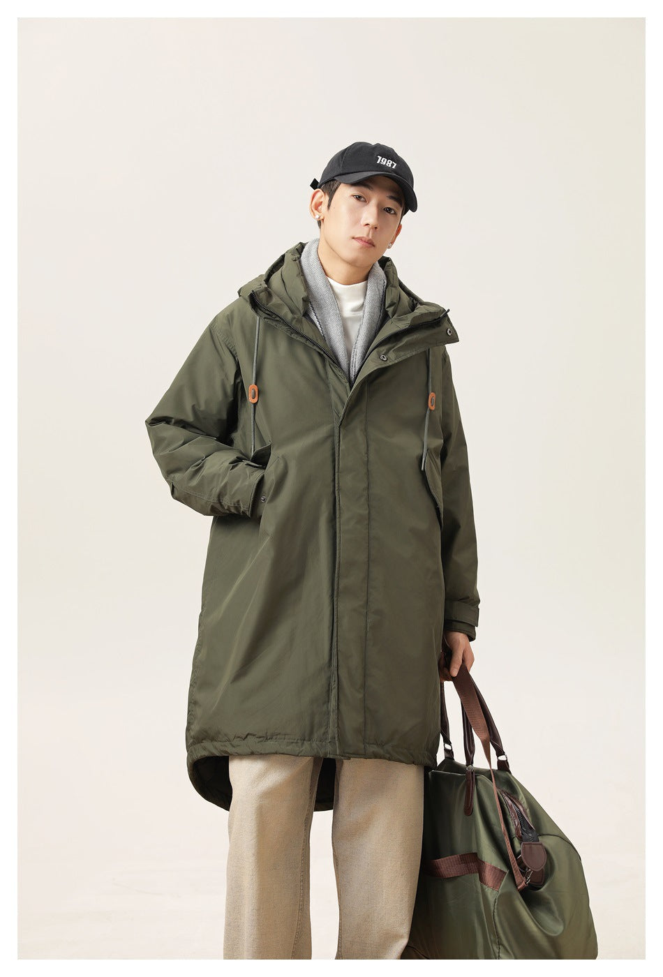 Model wearing an army green long hooded jacket over a gray hoodie layer, holding a green duffel bag