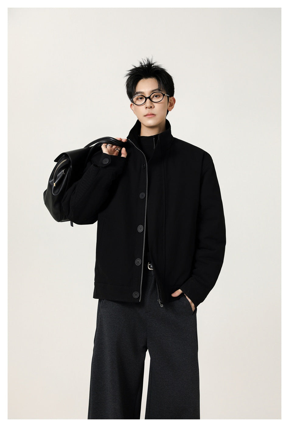 Model wearing a black high-collar jacket with a buttoned front and zipper, carrying a black shoulder bag.
