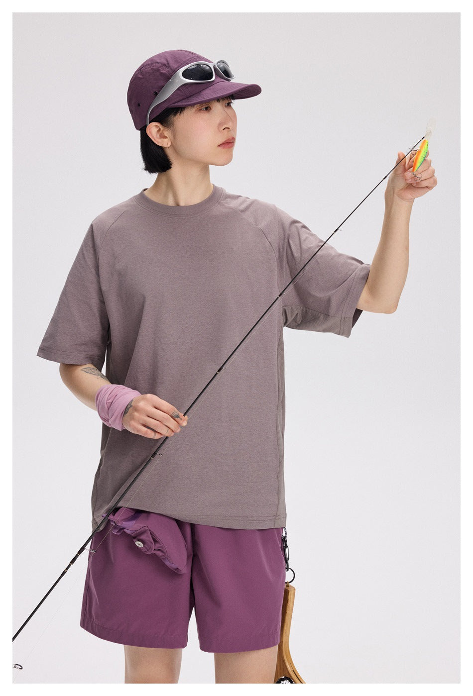 Model wearing a mauve oversized T-shirt and purple shorts, with a purple cap and sunglasses, holding a fishing rod.