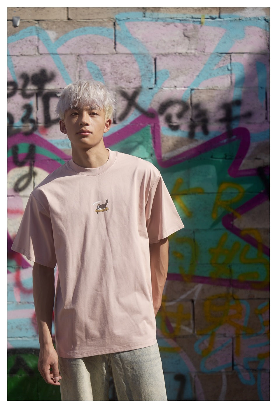 Model wearing a pale pink oversized T-shirt with a small cat-on-skateboard chest graphic, facing the camera.