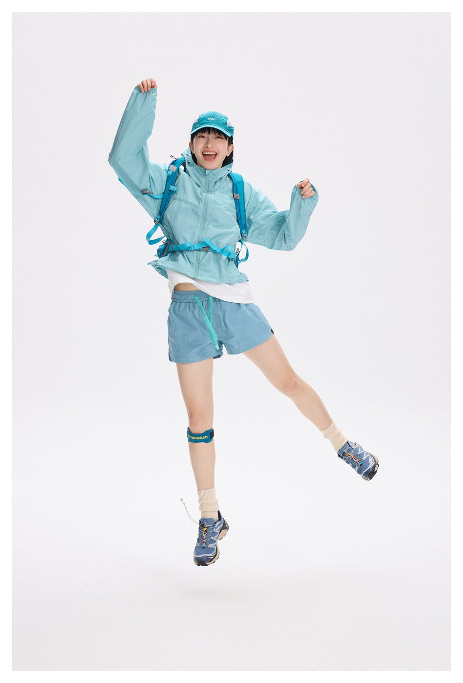 Model jumping in a light turquoise hooded jacket and blue shorts with a hiking backpack.