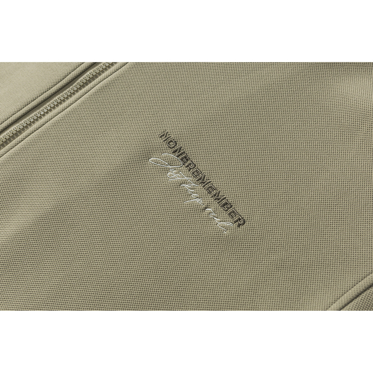 Close-up of embroidered lettering on olive jacket fabric beside the zipper.