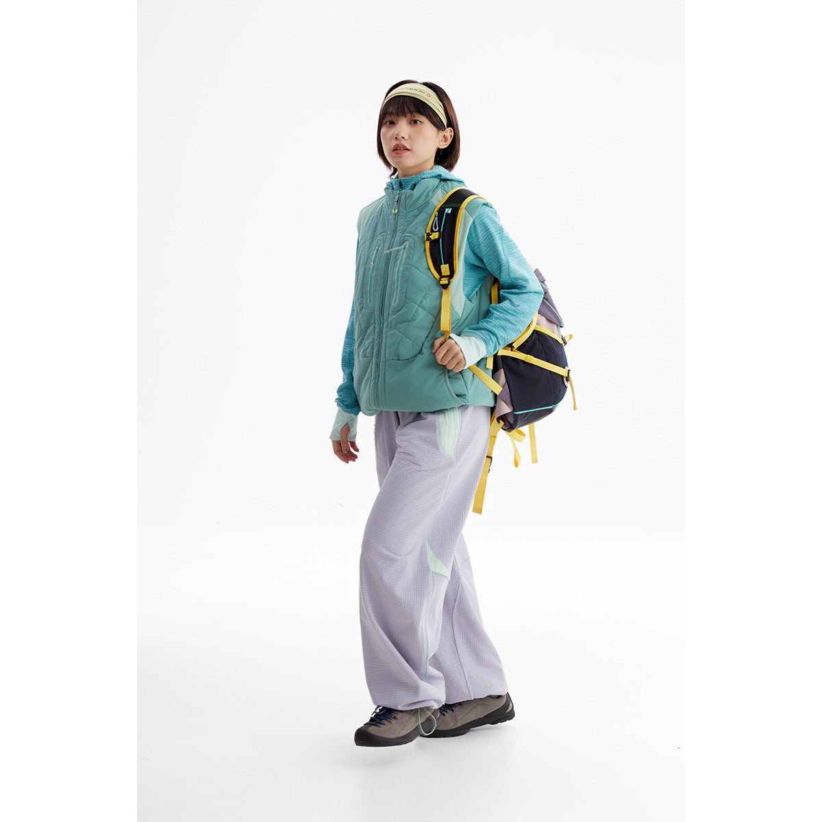 Full-length model wearing a teal quilted vest and blue hooded top with loose lavender pants and a hiking backpack.