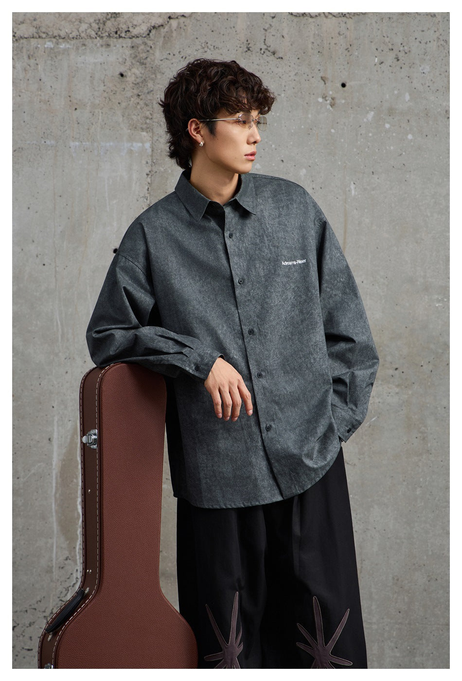 Model wearing an oversized charcoal gray button-up shirt with a small white chest print, posed beside a guitar case.