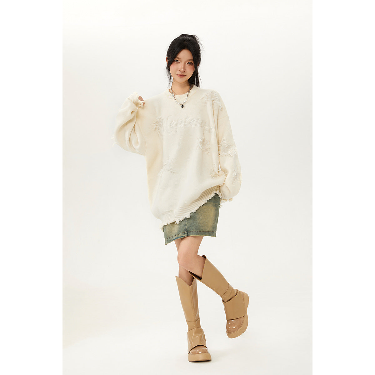 Model wearing an oversized cream sweater with distressed patches and script lettering, paired with a denim skirt and tan