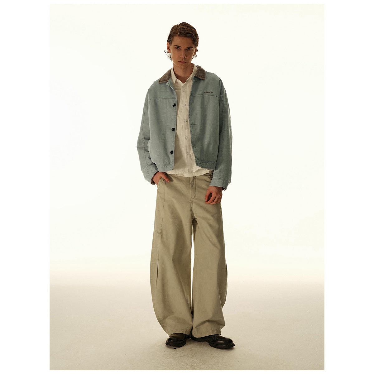 Model wearing a pale blue-gray button-front jacket with a contrasting brown collar and loose beige trousers.