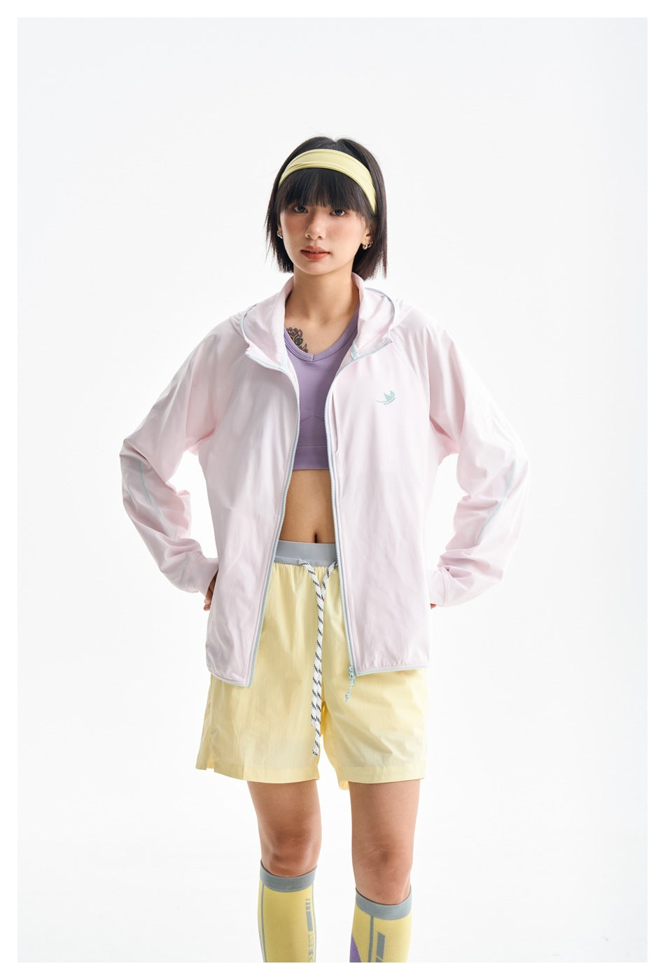 Closer front view of a model wearing a pale pink hooded jacket with aqua trim and yellow shorts.