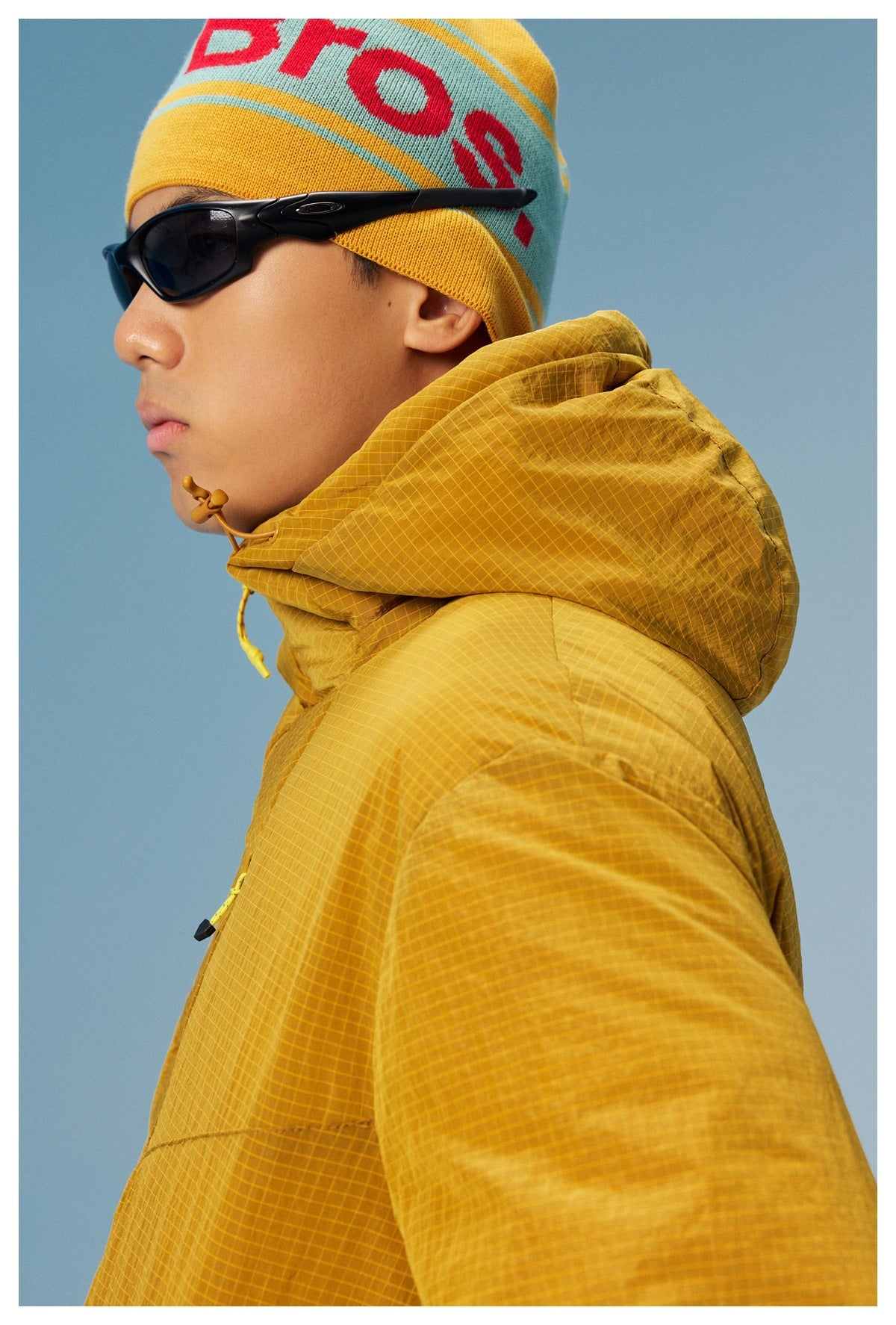 Close side view of a mustard grid-pattern pullover's hood, drawcord, and upper sleeve worn by a model.