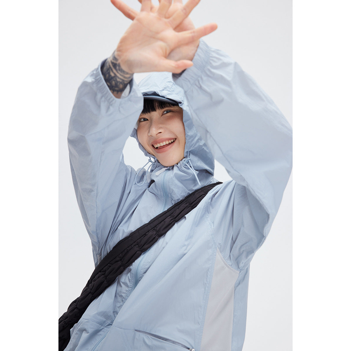 Model wearing a pale blue hooded jacket with a front zip and black crossbody strap, raising both arms.