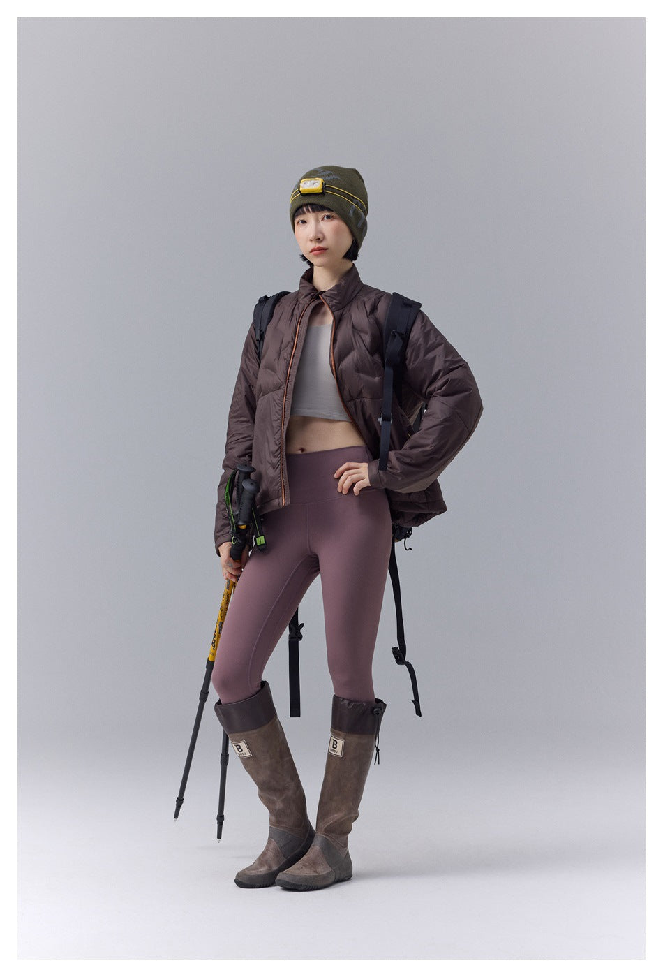 Full-length model wears a dark brown quilted jacket, mauve leggings, tall brown boots, and an olive beanie.