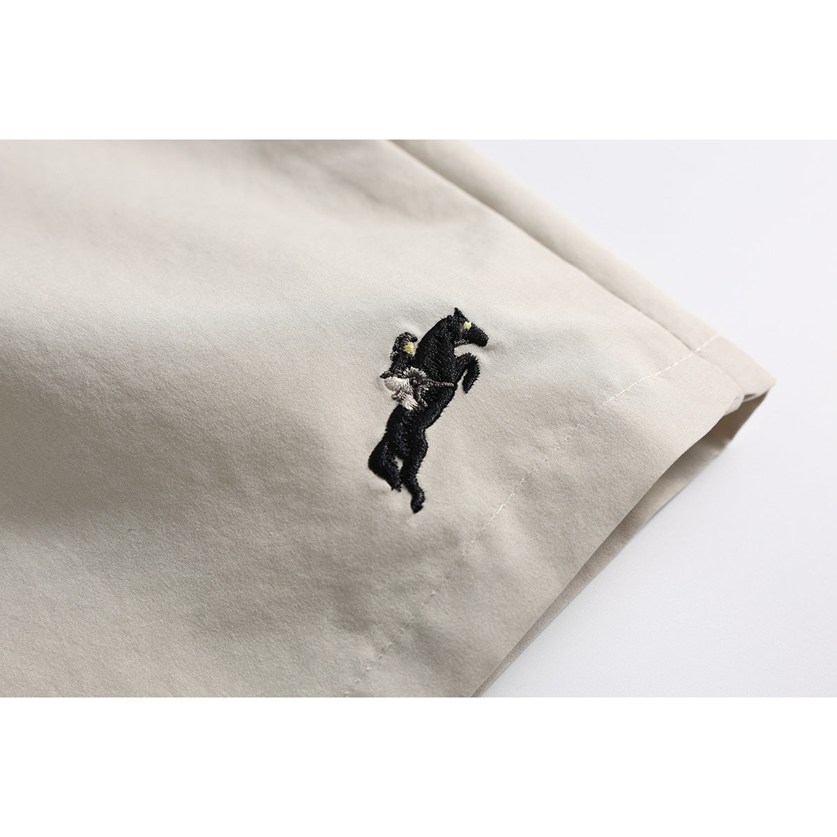 Close-up of beige shorts showing a small embroidered horse emblem near the hem.