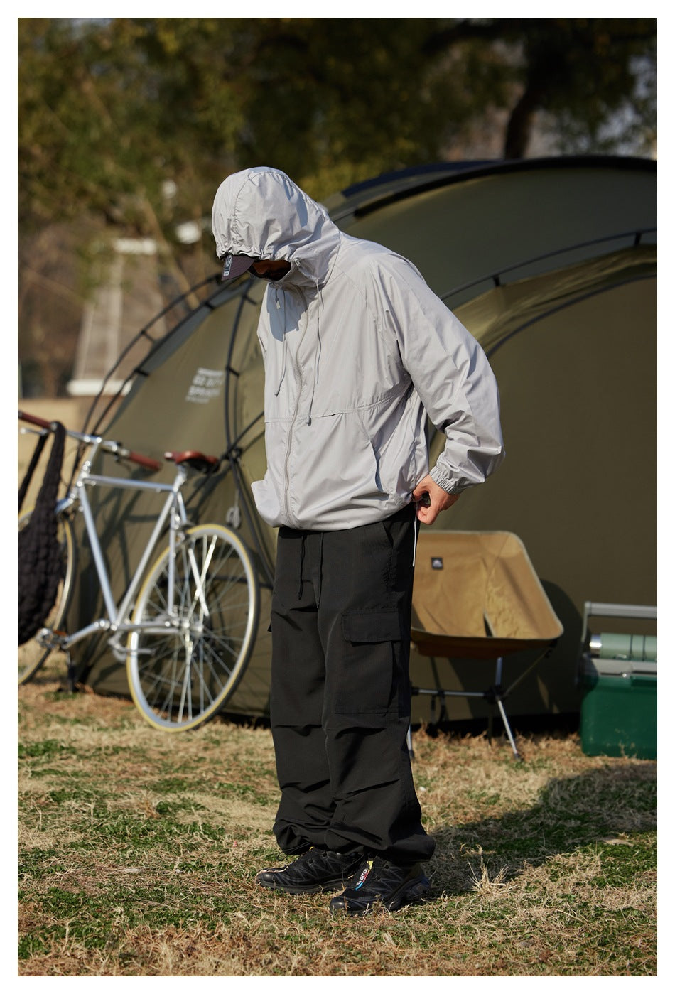 Model wearing a light gray hooded zip jacket and black cargo pants outdoors beside a tent.